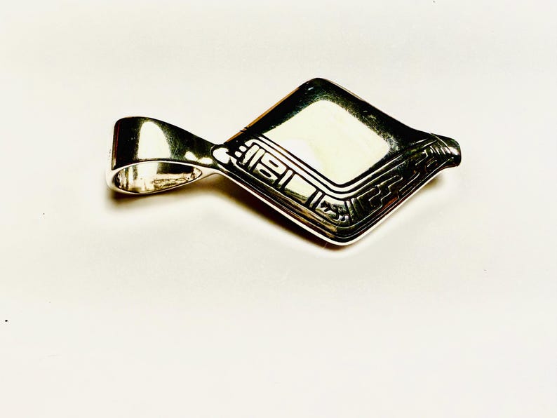 May include: A silver pendant with a geometric design. The pendant has a rectangular shape with a loop for a chain. The surface is decorated with engraved patterns and lines.