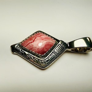 May include: A silver pendant with a rhombus-shaped pink gemstone. The gemstone has a marbled appearance with red and white hues. The pendant has an ornate, geometric design around the stone and a loop for a chain.