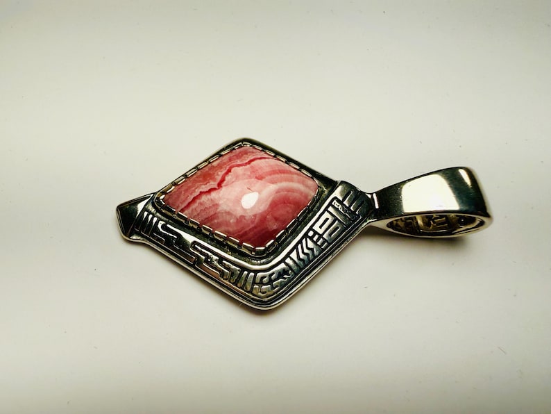 May include: A silver pendant with a rhombus-shaped pink gemstone. The gemstone has a marbled appearance with red and white hues. The pendant has an ornate, geometric design around the stone and a loop for a chain.