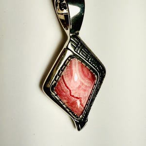 May include: A silver pendant with a rhodonite gemstone. The pendant has a geometric design around the pink and white stone. The bail is a simple loop. The pendant is displayed on a white background.