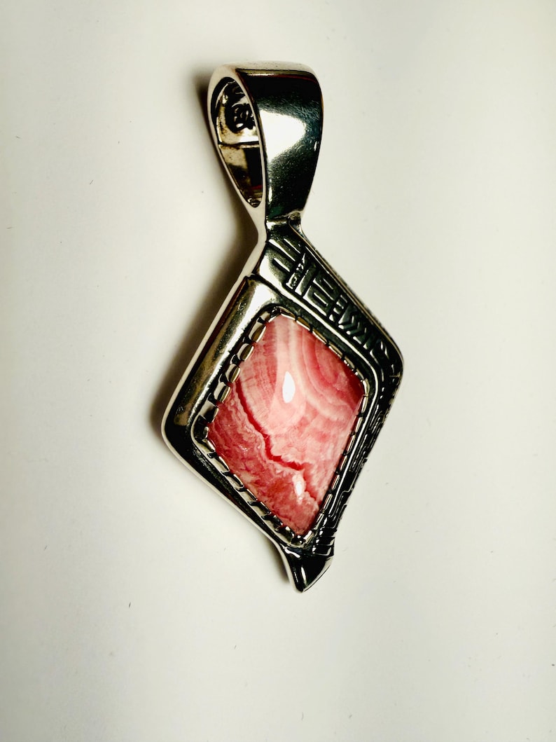 May include: A silver pendant with a rhodonite gemstone. The pendant has a geometric design around the pink and white stone. The bail is a simple loop. The pendant is displayed on a white background.