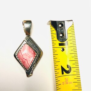 May include: A silver pendant with a pink and white stone in a diamond shape. The pendant is approximately 2 inches long, as measured by a yellow tape measure. The pendant has a decorative design around the stone.