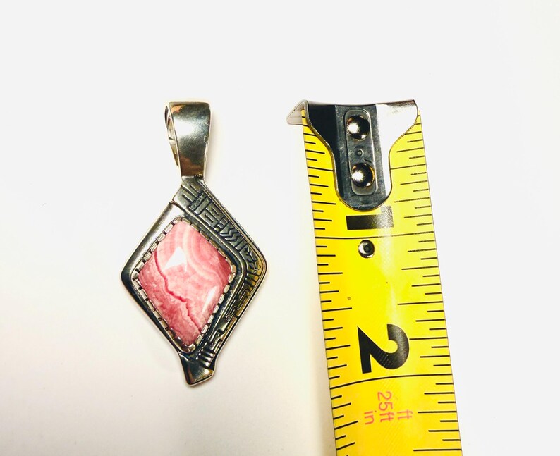 May include: A silver pendant with a pink and white stone in a diamond shape. The pendant is approximately 2 inches long, as measured by a yellow tape measure. The pendant has a decorative design around the stone.