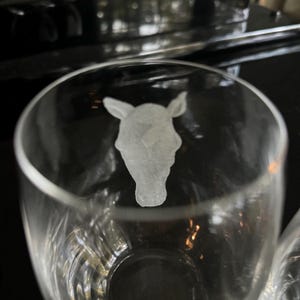 Puede incluir: Un vaso de vidrio transparente con un dise&ntilde;o grabado de una cabeza de caballo. El vaso est&aacute; vac&iacute;o y parece ser parte de un juego.