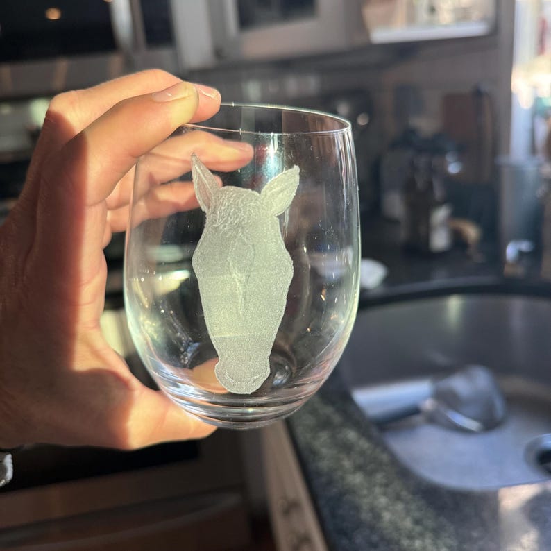 Puede incluir: Una copa de vino de vidrio transparente con una cabeza de caballo blanca grabada en el lateral.
