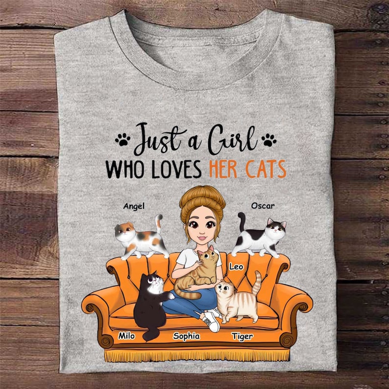 Puede incluir: Una camiseta gris con el texto "Just a Girl Who Loves Her Cats" y una ilustraci&oacute;n de dibujos animados de una mujer sentada en un sof&aacute; con cinco gatos. Los gatos se llaman Angel, Oscar, Leo, Milo y Tiger.