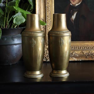May include: Two antique brass cocktail shakers with ornate detailing. The shakers are cylindrical with a flared base and a decorative band near the top. They sit on a dark surface, with a framed portrait and a lit candle in the background.
