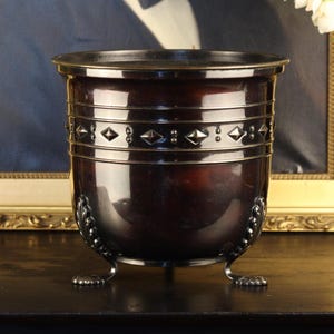 May include: A dark metallic planter with a smooth, rounded body and a wide rim. The upper section features a horizontal band decorated with raised diamond shapes and small dots. It stands on three curved, ornate feet with textured details.
