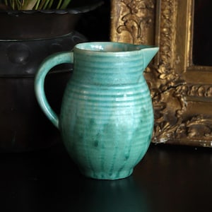 May include: A teal blue ceramic jug with a rounded body and a handle. The jug has a glazed finish with a subtle striped pattern.