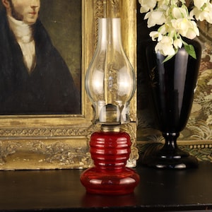 May include: A vintage-style oil lamp featuring a deep red, horizontally ribbed glass base and a clear, bulbous glass chimney. The lamp has a metallic burner assembly connecting the red glass reservoir to the transparent glass top section.