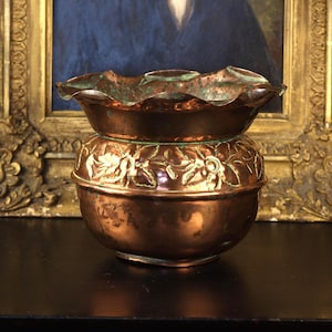May include: A copper planter with a wide, ruffled rim and a rounded body. The exterior features a raised floral relief pattern around the centre. The metal surface displays a warm, polished copper tone with visible green oxidation in the crevices.