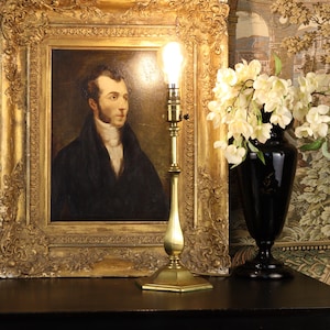 May include: A portrait in a gold ornate frame is lit by a brass lamp. A black vase holds white flowers. An antique green glass lamp is on the left. The background features a tapestry with a landscape design. The scene is set on a dark wooden surface.