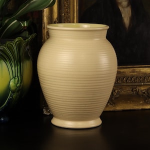 May include: A cream-colored, ribbed ceramic vase with a rounded body and a slightly flared rim. The vase sits on a small, circular base. The background includes a framed portrait and a green and yellow decorative planter.