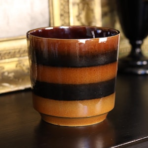 May include: A cylindrical ceramic container with a layered design of brown, black, and tan bands. The interior is a darker brown. The container sits on a dark surface, with a gold-framed object and a black vase in the background.