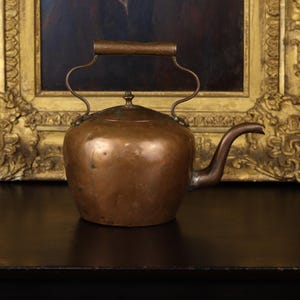 May include: A vintage copper kettle with a curved spout and a handle. The kettle has a rounded body and a lid with a small knob. The handle is made of a darker material. The kettle is sitting on a dark surface.