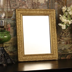 May include: A gold-colored framed mirror with ornate detailing. A green glass lamp with a black base is to the left. A black vase with white flowers is to the right. The items are on a dark surface.