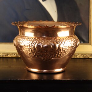 May include: A polished copper planter featuring a scalloped rim and a rounded body. The exterior surface displays a detailed hammered texture combined with embossed floral motifs that wrap around the centre of the vessel in a repeating pattern.
