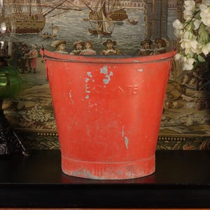 May include: A weathered, red metal bucket with a slightly tapered shape. The bucket has a metal rim and a faded appearance, with visible signs of wear and tear. The number "76" is visible on the side. The bucket is an antique decorative item.