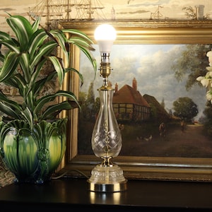 May include: A clear glass table lamp with a diamond pattern, brass accents, and a white bulb. The lamp sits on a black surface, with a framed painting and a green potted plant in the background.