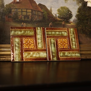 May include: Two square ceramic tiles with a geometric design. The tiles feature a pattern of brown, green, and gold, with a central floral motif. The tiles are displayed on a dark surface, with a framed painting in the background.