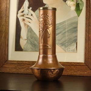 May include: A cylindrical copper vase with a flared base and a metallic rim. The body features an embossed floral design with stems, leaves, and small berries. The copper surface has a warm, polished finish with subtle variations in tone.