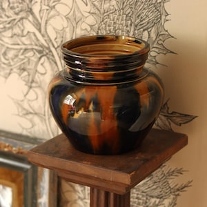 May include: A round ceramic pot featuring a glossy glaze with a marbled pattern of deep black and warm amber tones. The vessel has a wide, curved body that tapers toward a short, ridged neck with a circular opening at the top.