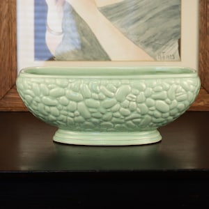 May include: A light green ceramic planter with a pebble-like texture. The oval-shaped planter sits on a small, round base. The background includes a framed artwork with a wooden frame.
