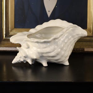 May include: A white ceramic seashell-shaped planter. The planter has a textured surface with a scalloped edge and sits on three small feet. The interior is smooth and light gray. The background includes a framed portrait.