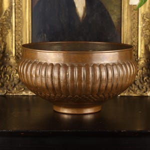May include: A round copper bowl featuring a wide, open top and a tapered base. The exterior surface is decorated with a series of vertical, rounded fluted ridges around the upper section and a textured, cross-hatched pattern near the bottom.