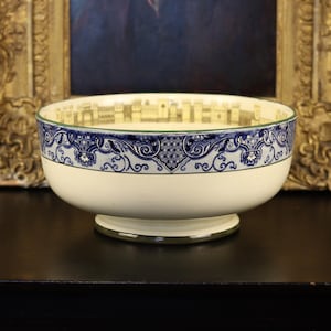 May include: A cream-colored ceramic bowl with a blue and white decorative border. The bowl has a footed base and a green rim. The interior features a cityscape design. The bowl is likely vintage or antique.