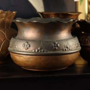 May include: A copper bowl with a scalloped rim and a dark, oxidised band around the centre featuring embossed floral motifs and dotted patterns. The lower portion of the bowl has a smooth, rounded shape with a warm, metallic finish.