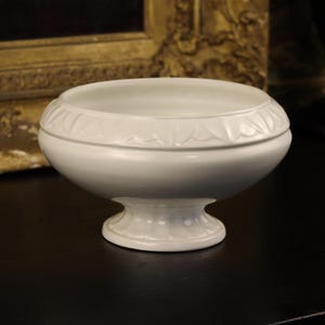May include: A white, pedestal bowl with a floral pattern around the rim. The bowl has a rounded shape and sits on a short, flared base. The background includes a framed artwork and a dark surface.
