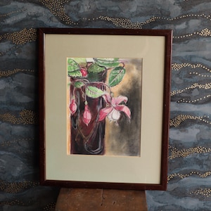 May include: A framed watercolour painting of a fuchsia plant with pink and white flowers, green leaves, and red buds. The painting is in a dark brown frame with a cream-coloured mount. The background is a dark, textured wall.