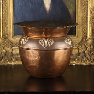 May include: A round copper planter with a scalloped rim and a series of embossed shell-like motifs encircling the upper section. The metal surface displays a warm, metallic finish with subtle variations in texture and tone across the body.