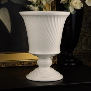 May include: A white ceramic pedestal vase featuring a flared, scalloped rim and a body decorated with diagonal, swirling fluted patterns. The vase stands on a circular, matching base with a similar spiraling texture, creating a cohesive design.