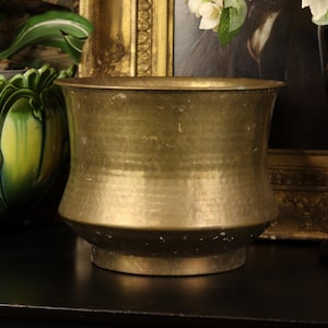 May include: A hammered brass planter with a wide, flared rim and a slightly tapered base. The planter has a warm, golden hue and a textured surface. It is sitting on a dark surface, with other decorative items in the background.