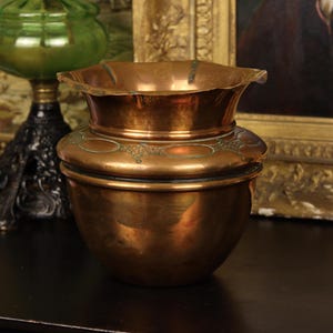 May include: A copper-colored antique vase with a flared rim and decorative etched details. The vase has a bulbous base and a slightly curved body. The background includes a green glass lamp and a framed painting.