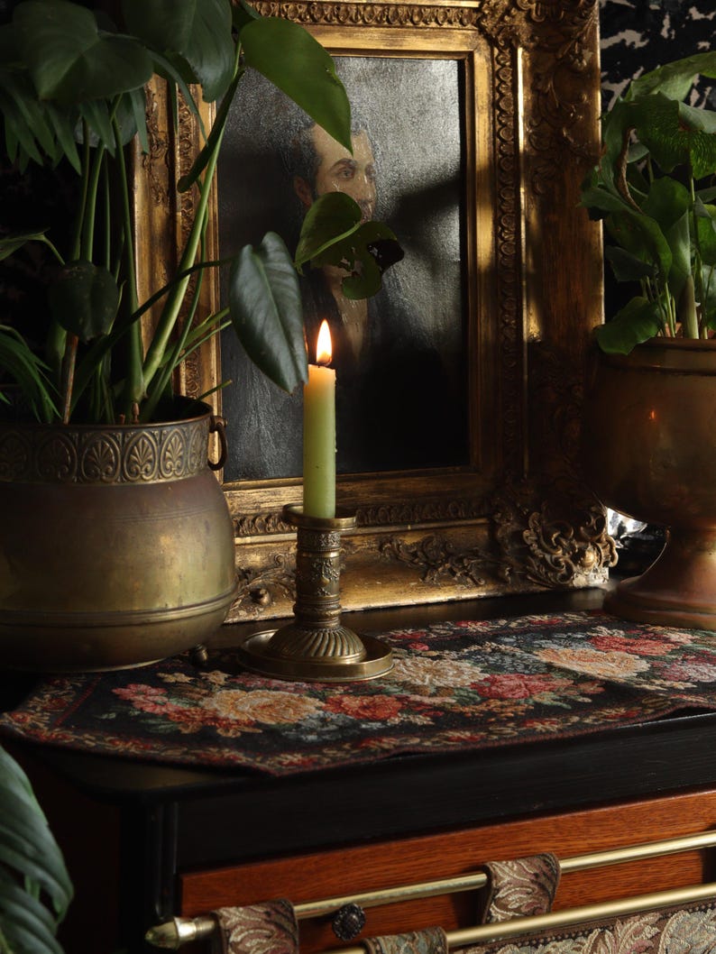 May include: A lit green candle in a brass candlestick sits on a black and floral patterned table runner. A brass planter with a green plant is in the background.