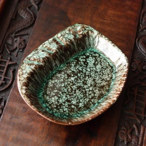 May include: A ceramic, leaf-shaped dish with a speckled, teal and brown glaze. The dish has a glossy finish and sits on a dark wooden surface. The rim of the dish is a lighter color, creating a contrast with the darker interior.