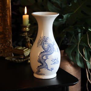 May include: A white ceramic vase featuring a blue dragon motif. The vase has a bulbous body and a flared neck. The dragon is intricately illustrated, encircling the vase. The vase is placed on a dark surface, with a candle and foliage in the background.