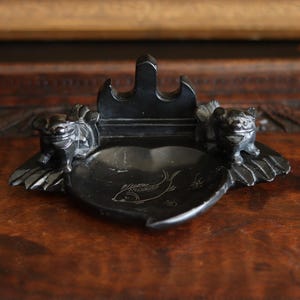 May include: A black, heart-shaped ashtray with two sculpted lion-like figures on either side. The ashtray features a central well with a fish design etched into the surface. The piece is made of a dark, possibly stone-like material.