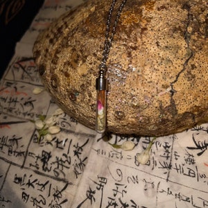 May include: A clear glass vial pendant necklace filled with dried flowers and herbs, suspended from a silver chain. The pendant is displayed against a textured, light brown background. Scattered jasmine flowers and script paper add to the aesthetic.
