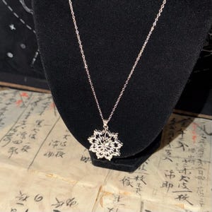 May include: A silver-toned necklace featuring a delicate chain and a pendant shaped like a multi-layered, symmetrical floral or star-like geometric pattern with intricate cut-out details that create a reflective, textured appearance.