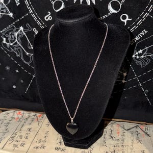May include: A silver chain necklace with a black heart-shaped pendant displayed on a black mannequin. The background features a black cloth with white astrological symbols and a vintage paper with Japanese characters.