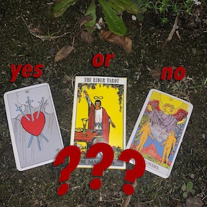 Op de afbeelding: Een tarotkaart lay-out met de rode woorden "yes" en "no". Drie tarotkaarten worden weergegeven: "Drie Zwaarden", "De Magiër" en "De Geliefden". Rode vraagtekens zijn voor de kaarten geplaatst. De Rider Tarot-doos is ook zichtbaar.