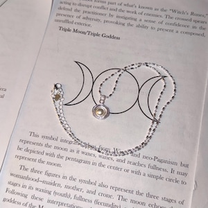 May include: A silver-toned necklace featuring a small, circular pendant with a central cream-colored pearl-like bead. The chain is composed of small, connected metallic beads and includes an adjustable extender chain with a tiny heart-shaped charm.