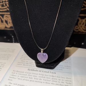 Op de afbeelding: Een hartvormige, lichtpaarse edelstenen hanger aan een dunne zwarte koordketting. De gladde, gepolijste steen is met een zilverkleurig metalen oogje aan het koord bevestigd, wat zorgt voor een eenvoudig en strak sieraad.