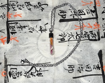 Peut inclure: Un collier argenté avec un petit pendentif en forme de flacon en verre rempli de fleurs séchées. Le flacon a un bouchon noir et est suspendu à une chaîne délicate. Le fond est un papier texturé avec de la calligraphie asiatique et des accents orange.
