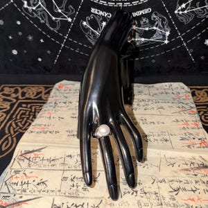 May include: A silver ring featuring a large, round, white pearl is shown on a black hand. The ring is set against a backdrop of a black cloth with white zodiac symbols and a piece of paper with handwritten text.