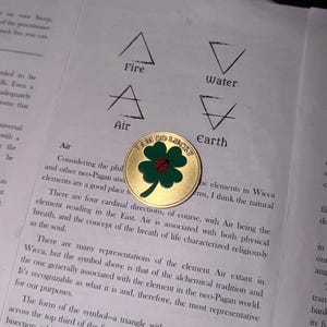 May include: A round gold-toned metal coin featuring a green four-leaf clover with a small red ladybug in the center. The text I AM SO LUCKY is embossed along the top edge of the coin, which is placed on a page with printed text and symbols.